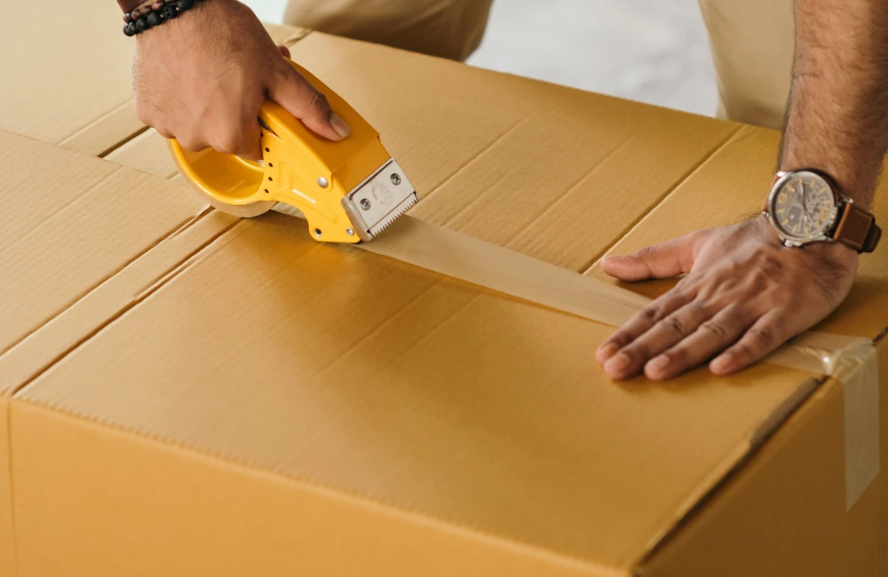 Close-up of a person using a yellow tape dispenser gun to seal a brown cardboard box for shipment.
